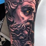 Este tatuaje neotradicional representa a Zeus, el dios griego. La pieza destaca por sus detalles en el cabello y la barba, utilizando sombras para crear profundidad. El contraste entre los colores oscuros y claros resalta los rasgos faciales, mientras que el toque dorado en el ojo añade un punto de enfoque. Las líneas fluidas del cabello y la barba aportan movimiento, y la composición general del tatuaje está bien equilibrada.